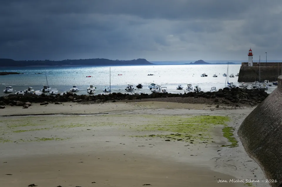 Bay at low tide under a stormy sky, boats moored in a silvery sea, red lighthouse on the jetty.